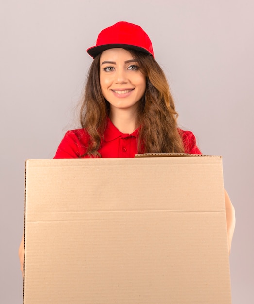 Jonge leveringsvrouw die rood poloshirt en GLB dragen die zich met kartondoos bevinden die vrolijk over geïsoleerde witte achtergrond glimlacht