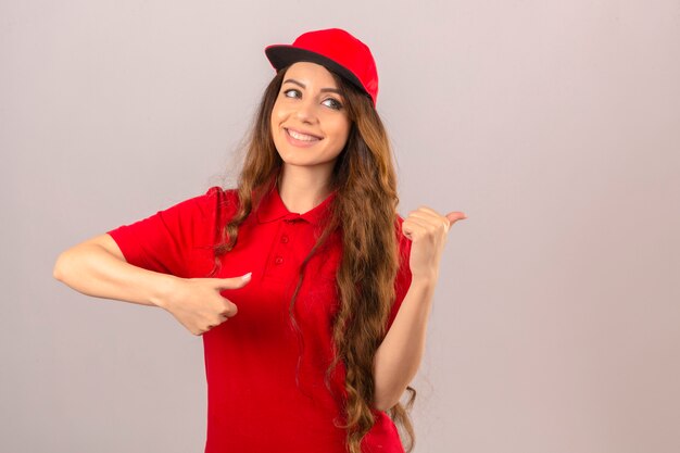 Jonge leveringsvrouw die rood poloshirt en pet draagt die met gelukkig gezicht glimlacht en naar de kant kijkt met duim omhoog over geïsoleerde witte achtergrond