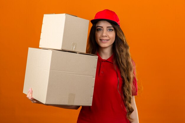 Jonge leveringsvrouw die rood poloshirt en pet draagt die zich met kartondozen bevinden die over geïsoleerde oranje achtergrond zelfverzekerd kijken