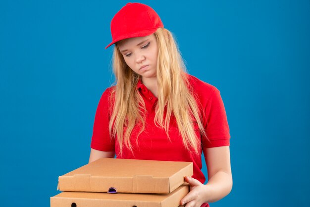 Jonge leveringsvrouw die rood poloshirt en pet draagt die zich met pizzadozen bevinden die verdrietig en verveeld kijken met een ongelukkig gezicht over geïsoleerde blauwe achtergrond