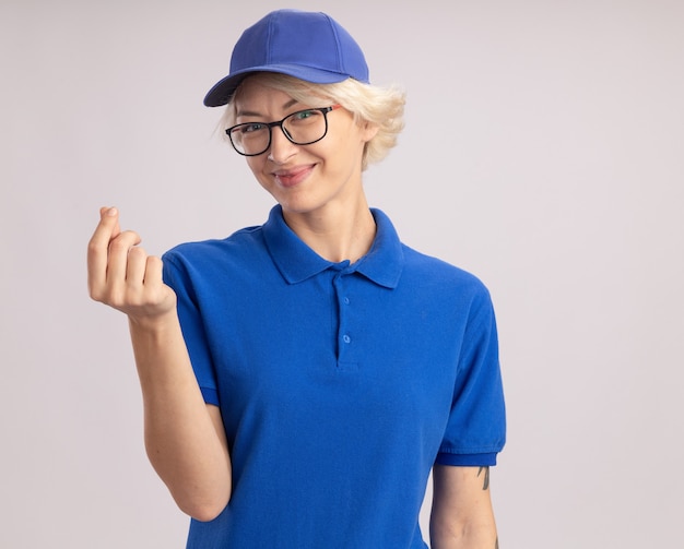 Jonge leveringsvrouw in blauw uniform en GLB die glazen dragen die het glimlachen wrijven vingers kijken die geldgebaar maken die zich over witte muur bevinden