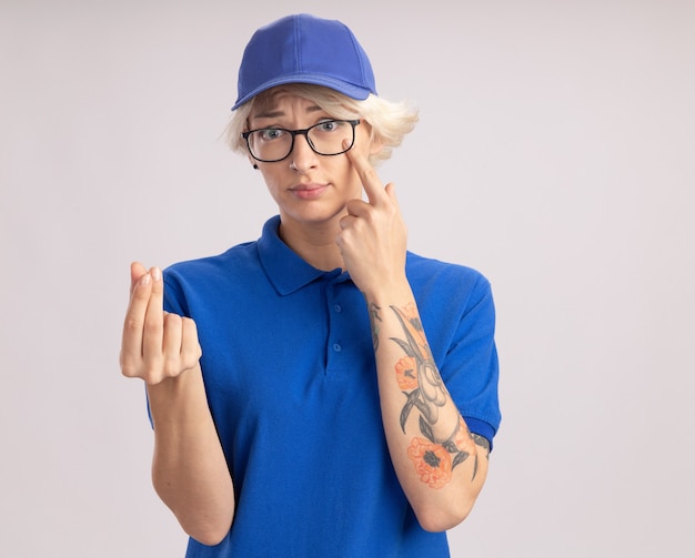 Jonge leveringsvrouw in blauw uniform en glb die glazen dragen die met vinger wijzen naar haar oog wrijven vingers die geldgebaar maken die zich over witte muur bevinden