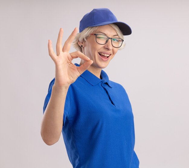 Jonge leveringsvrouw in blauw uniform en pet die knipogen en glimlachend tonend ok teken kijken dat zich over witte muur bevindt