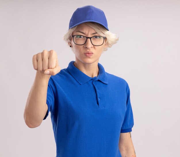 Jonge leveringsvrouw in blauw uniform en pet die met een boos gezicht kijken dat vuist toont die zich over witte muur bevindt