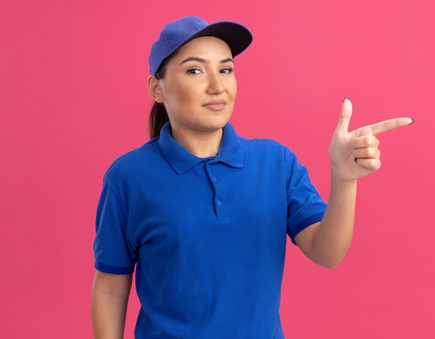 Jonge leveringsvrouw in blauw uniform en pet die naar voorzijde glimlachend zelfverzekerd kijken die met wijsvinger naar de kant over roze muur richt