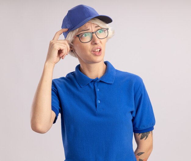 Jonge leveringsvrouw in blauw uniform en pet die opzij verbaasd krabend hoofd met vinger kijken die zich over witte muur bevindt