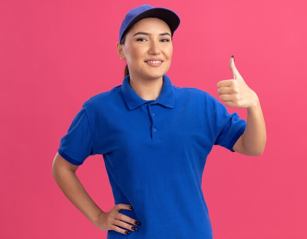 Jonge leveringsvrouw in blauw uniform en pet die voorzijde glimlachend zelfverzekerd zien zien thumbs up staande over roze muur kijken