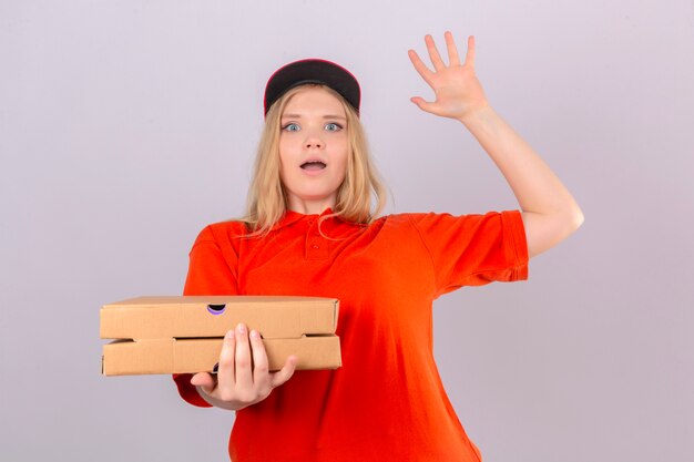 Jonge leveringsvrouw in oranje poloshirt en rode pet die verbaasd kijken terwijl hij mond open houdt over geïsoleerde witte achtergrond