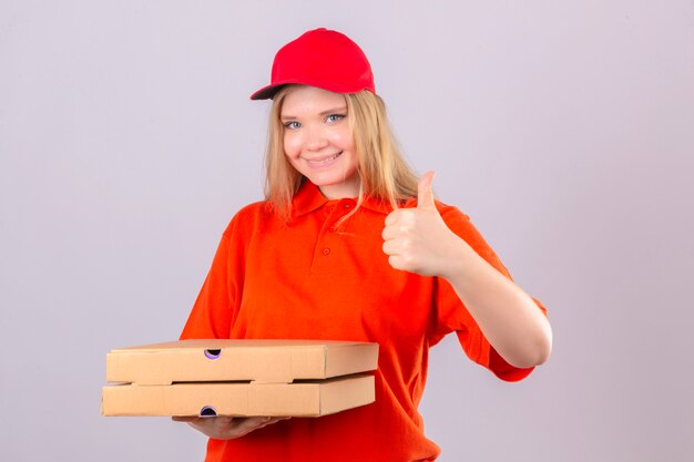 Jonge leveringsvrouw in oranje poloshirt en rode pet met pizzadozen die duim tonen die vrolijk over geïsoleerde witte achtergrond glimlachen