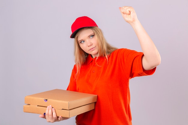 Jonge leveringsvrouw in oranje poloshirt en rode pet met pizzadozen die vuist opheffen na een concept van de overwinningswinnaar over geïsoleerde witte achtergrond