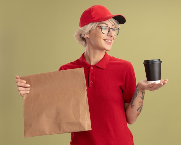 Jonge leveringsvrouw in rood uniform en GLB die glazen dragen die document pakket en document kop houden die zelfverzekerd glimlachend status over groene muur kijken