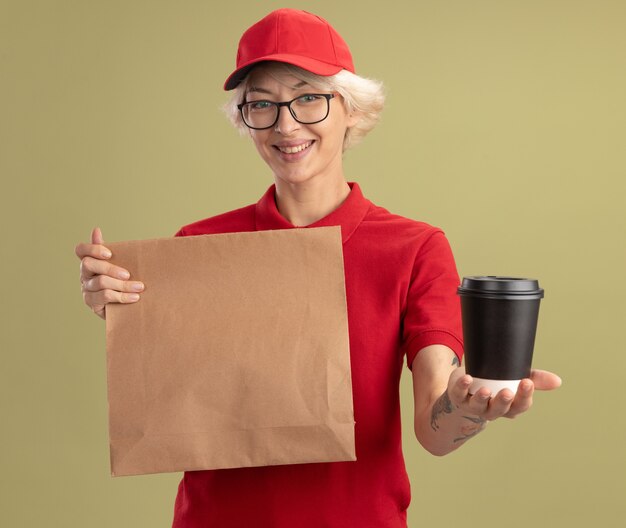 Jonge leveringsvrouw in rood uniform en GLB die glazen dragen die document pakket houden die koffiekop aanbieden die zich staande over groene muur glimlachen