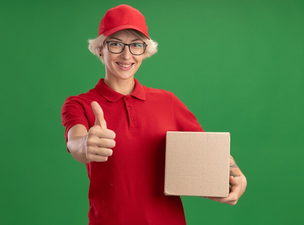 Jonge leveringsvrouw in rood uniform en GLB die glazen dragen die kartondoos houden die het tonen duimen die omhoog glimlachen vrolijk glimlachen die zich over groene muur bevinden