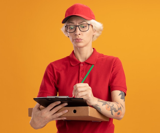 Jonge leveringsvrouw in rood uniform en GLB die glazen dragen die pizzadoos en klembord met potlood houden die iets met ernstig gezicht schrijven dat zich over oranje muur bevindt