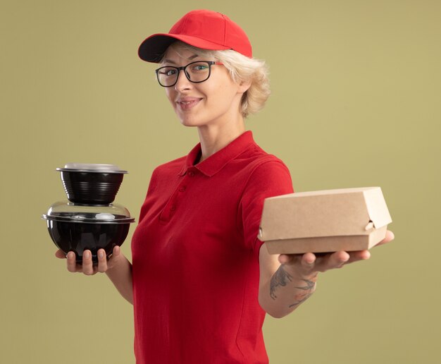 Jonge leveringsvrouw in rood uniform en GLB die glazen dragen die voedselpakketten smilign zelfverzekerd over groene muur houden