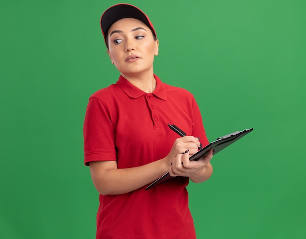 Jonge leveringsvrouw in rood uniform en GLB-holdingsklembord en potlood die opzij met ernstige zekere uitdrukking kijken die zich over groene muur bevindt