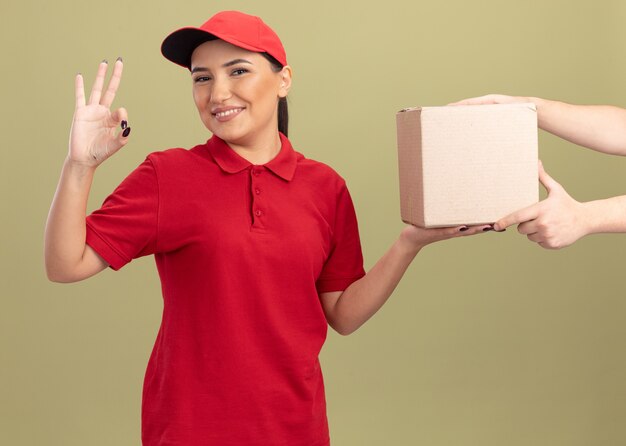 Jonge leveringsvrouw in rood uniform en pet die vriendelijk glimlachen tonend ok teken terwijl het ontvangen van doospakket zich over groene muur bevindt