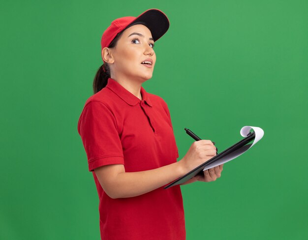 Jonge leveringsvrouw in rood uniform en pet met klembord en potlood opzoeken met peinzende uitdrukking denken staande over groene muur