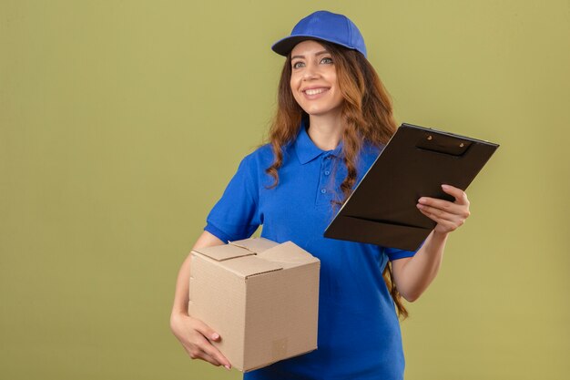 Jonge leveringsvrouw met krullend haar die blauw poloshirt en GLB dragen die zich met kartondoos en klembord bevinden die vriendschappelijk over geïsoleerde groene achtergrond glimlachen