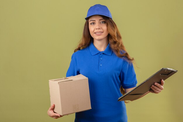 Jonge leveringsvrouw met krullend haar die blauw poloshirt en GLB dragen die zich met kartondoos en klembord bevinden die vriendschappelijk over geïsoleerde groene achtergrond glimlachen