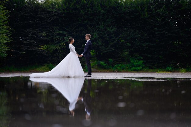 Jonge liefdevolle bruiden paar hand in hand staande in de buurt van meer kijken naar elkaar mooie bruid vrouw in gezwollen jurk met man man in elegant pak familie bruiloft dagviering