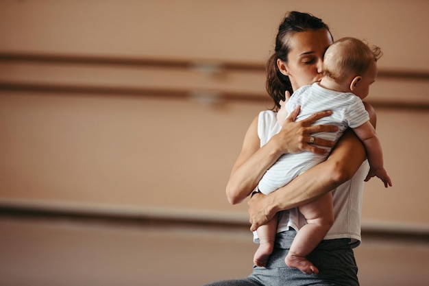 Gratis foto jonge liefhebbende moeder die haar baby kust terwijl ze hem vasthoudt tijdens de trainingsles in de gezondheidsclub ruimte kopiëren