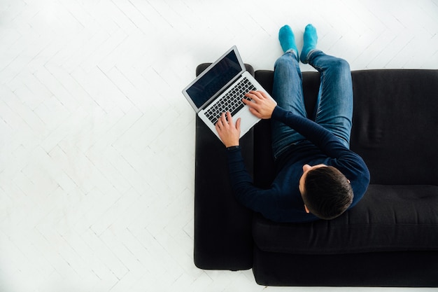 Jonge man aan het werk op laptop, zittend op een zwarte bank, witte vloer.