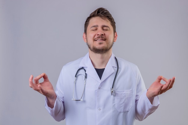 Jonge man arts dragen witte jas en stethoscoop ontspannen en lachend met de ogen gesloten doen meditatie gebaar met vingers