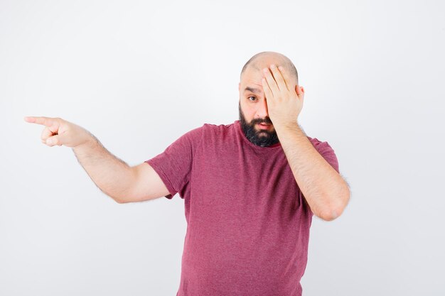 Jonge man bedekt zijn ene oog terwijl hij opzij wijst in een roze t-shirt en er verontrust uitziet. vooraanzicht.