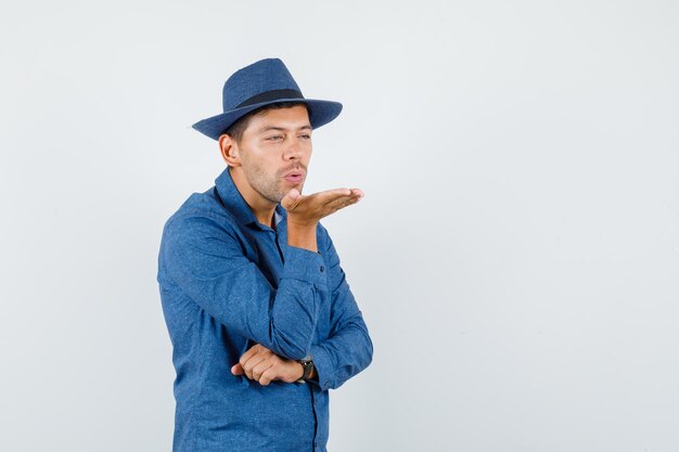 Jonge man blaast luchtkus met pruilende lippen in blauw shirt, hoed, vooraanzicht.