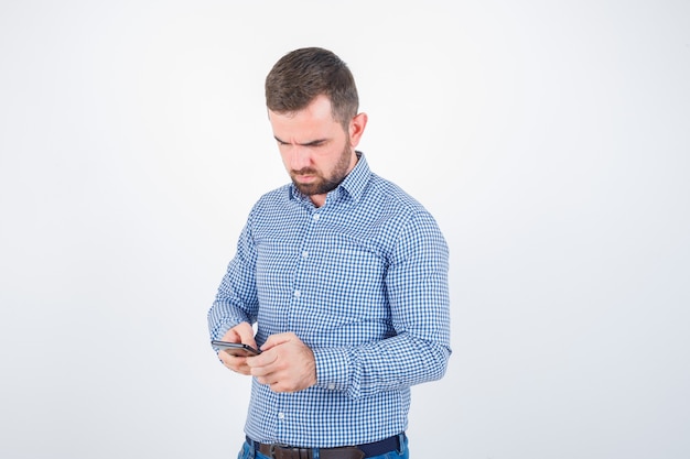 Jonge man chatten op mobiele telefoon in shirt, spijkerbroek en peinzend, vooraanzicht kijken.