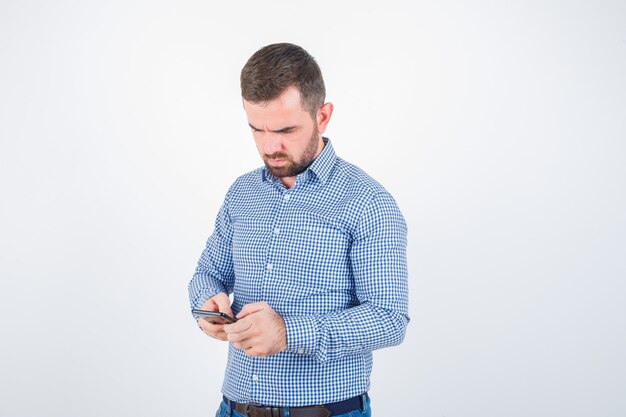 Jonge man chatten op mobiele telefoon in shirt, spijkerbroek en peinzend, vooraanzicht kijken.