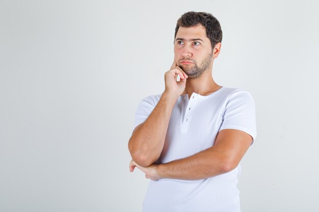 Jonge man denken met vinger op wang in wit t-shirt.