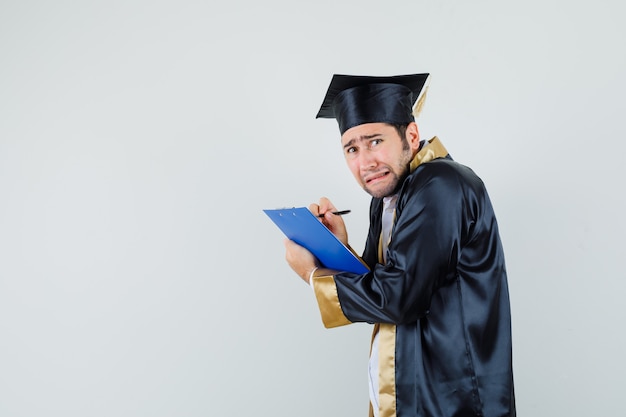 Jonge man die aantekeningen maakt op het klembord in afgestudeerde uniform en depressief kijkt.