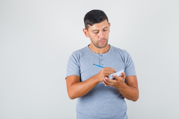 Jonge man die aantekeningen op notitieboekje in grijs t-shirt maakt en voorzichtig kijkt.