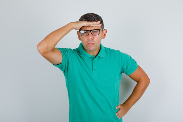 Jonge man die camera onderzoekt door hand over ogen in groen t-shirt, glazen te zetten.