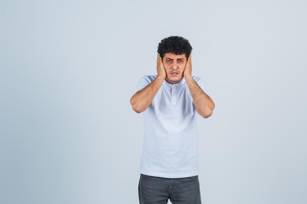 Jonge man die de handen op de oren drukt in een wit t-shirt en een spijkerbroek en er gehaast uitziet, vooraanzicht.