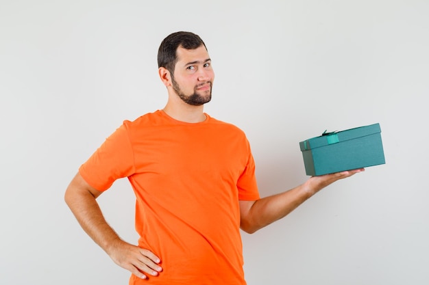 Jonge man die de huidige doos in een oranje t-shirt vasthoudt en er zelfverzekerd uitziet, vooraanzicht.