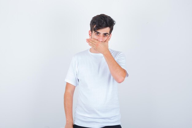 Jonge man die de mond bedekt met de hand in een t-shirt en er zelfverzekerd uitziet