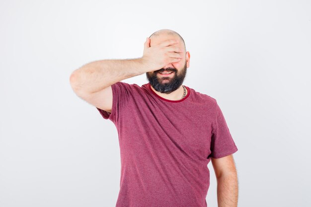 Jonge man die de ogen bedekt met de hand terwijl hij lacht in een roze t-shirt en er optimistisch uitziet, vooraanzicht.