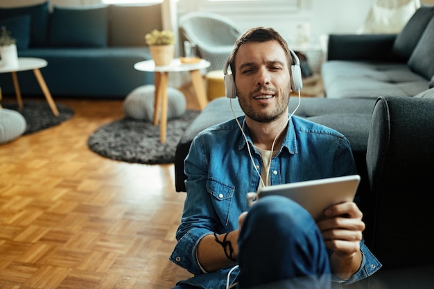 Jonge man die digitale tablet gebruikt en geniet van muziek via een koptelefoon terwijl hij ontspant in de woonkamer.