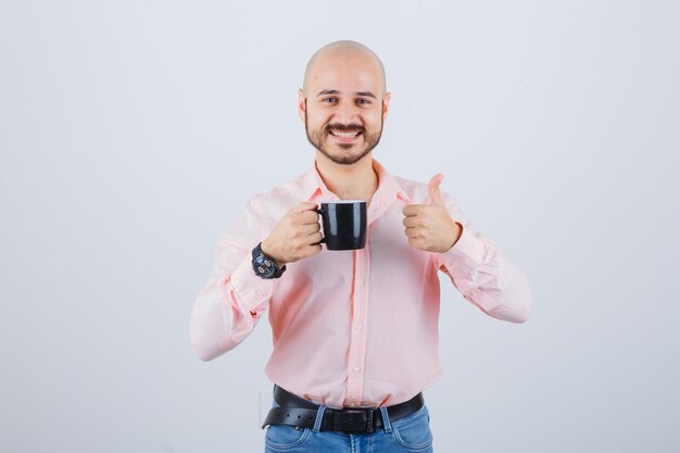 Jonge man die een beker vasthoudt terwijl hij zijn duim in een roze shirt, spijkerbroek laat zien en er vrolijk uitziet, vooraanzicht.