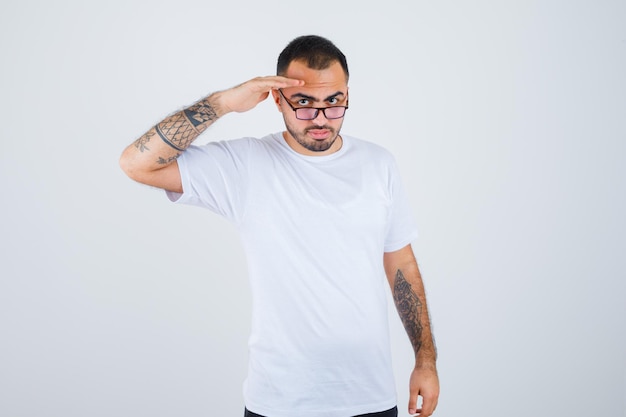 Jonge man die een bril draagt en een saluutgebaar toont in een wit t-shirt en een zwarte broek en er serieus uitziet