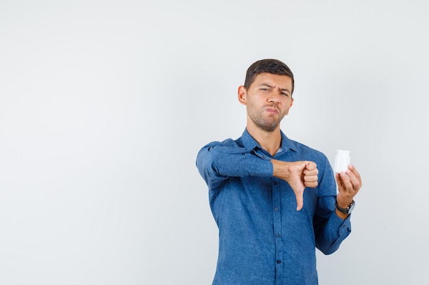 Jonge man die een fles pillen vasthoudt met duim naar beneden in een blauw shirt en er ontevreden uitziet. vooraanzicht.