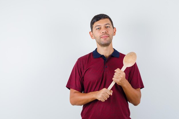 Jonge man die een houten lepel in een t-shirt houdt en er vrolijk uitziet. vooraanzicht.