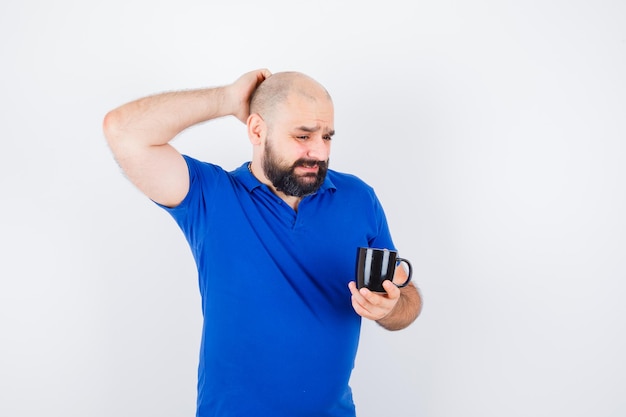 Jonge man die een kop vasthoudt terwijl hij zijn hoofd krabt in het vooraanzicht van het blauwe shirt.