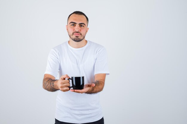 Jonge man die een kopje thee presenteert in een wit t-shirt en een zwarte broek en er serieus uitziet