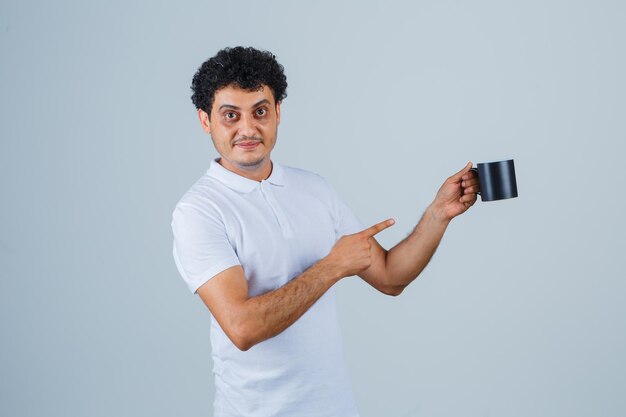 Jonge man die een kopje thee vasthoudt, ernaar wijst in een wit t-shirt en een spijkerbroek en er gelukkig uitziet. vooraanzicht.