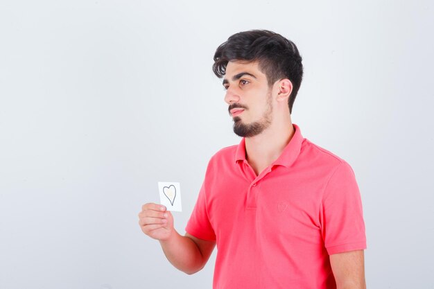 Jonge man die een plakbriefje vasthoudt terwijl hij wegkijkt in een t-shirt en er attent uitziet, vooraanzicht.