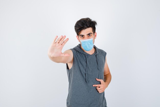 Jonge man die een stopgebaar toont terwijl hij een masker in een grijs t-shirt draagt en er serieus uitziet